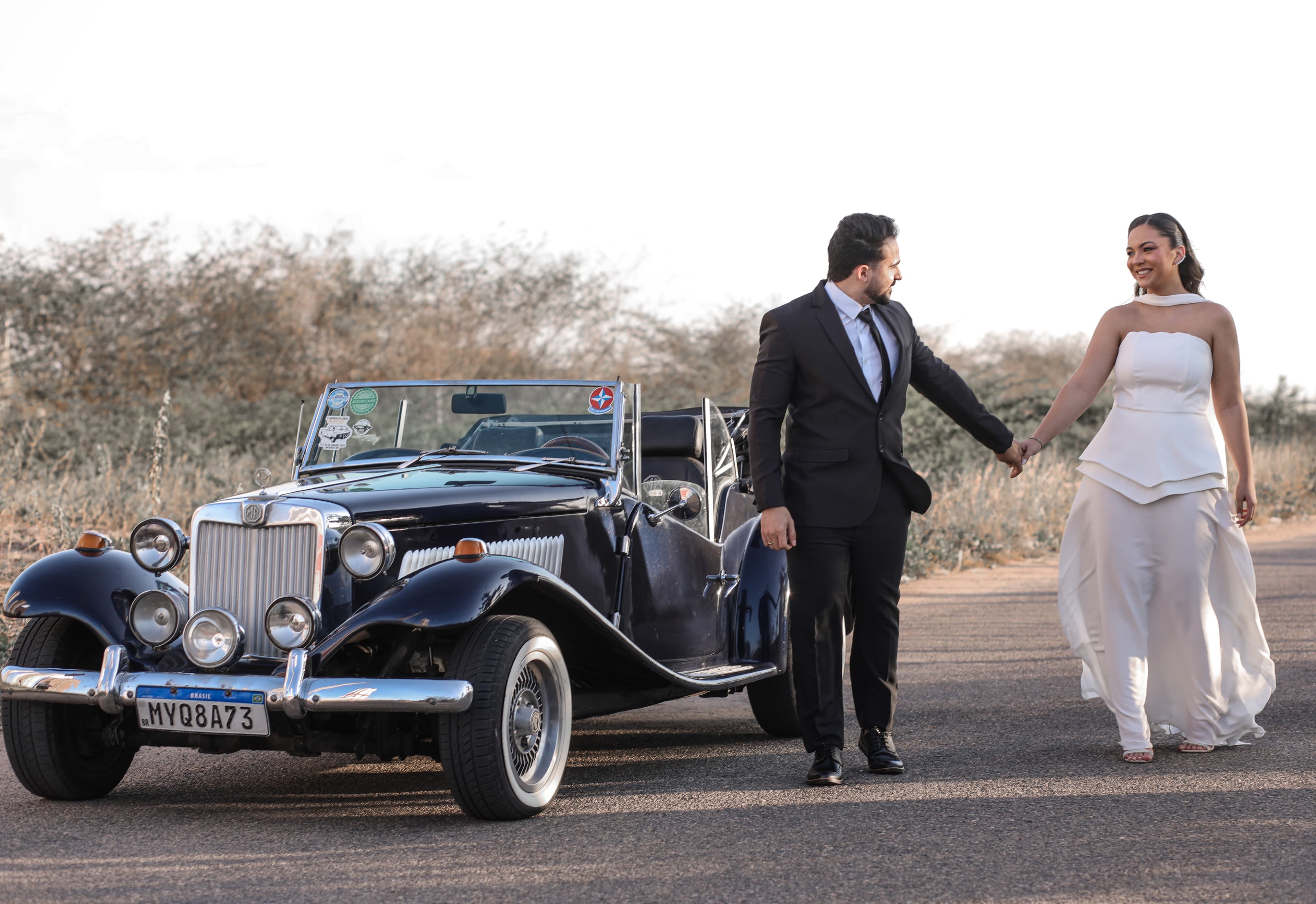 Rellyson pedido a mão de Gabrielly enquanto estão caminhando ao lado do carro MG azul.